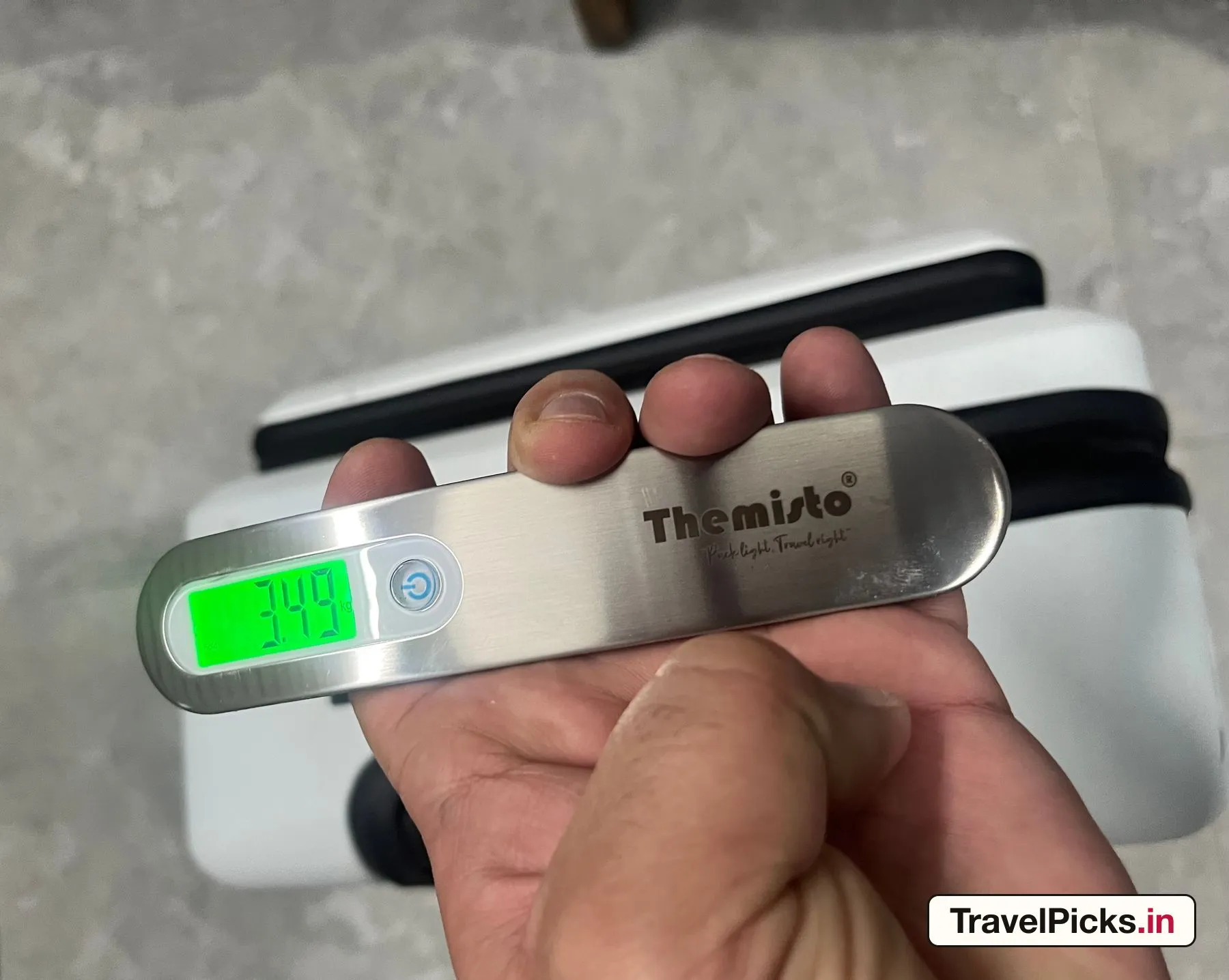 A digital luggage scale held in front of the case, its lit display reading 3.49 kg.