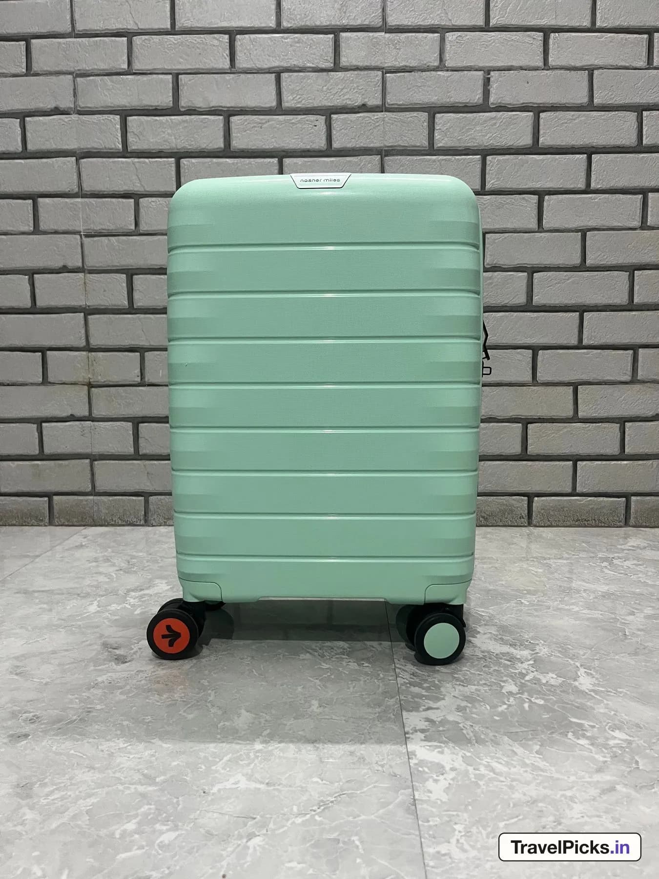 The mint green case standing upright against a white brick wall, handle down, showing its horizontal ribs and four double wheels.