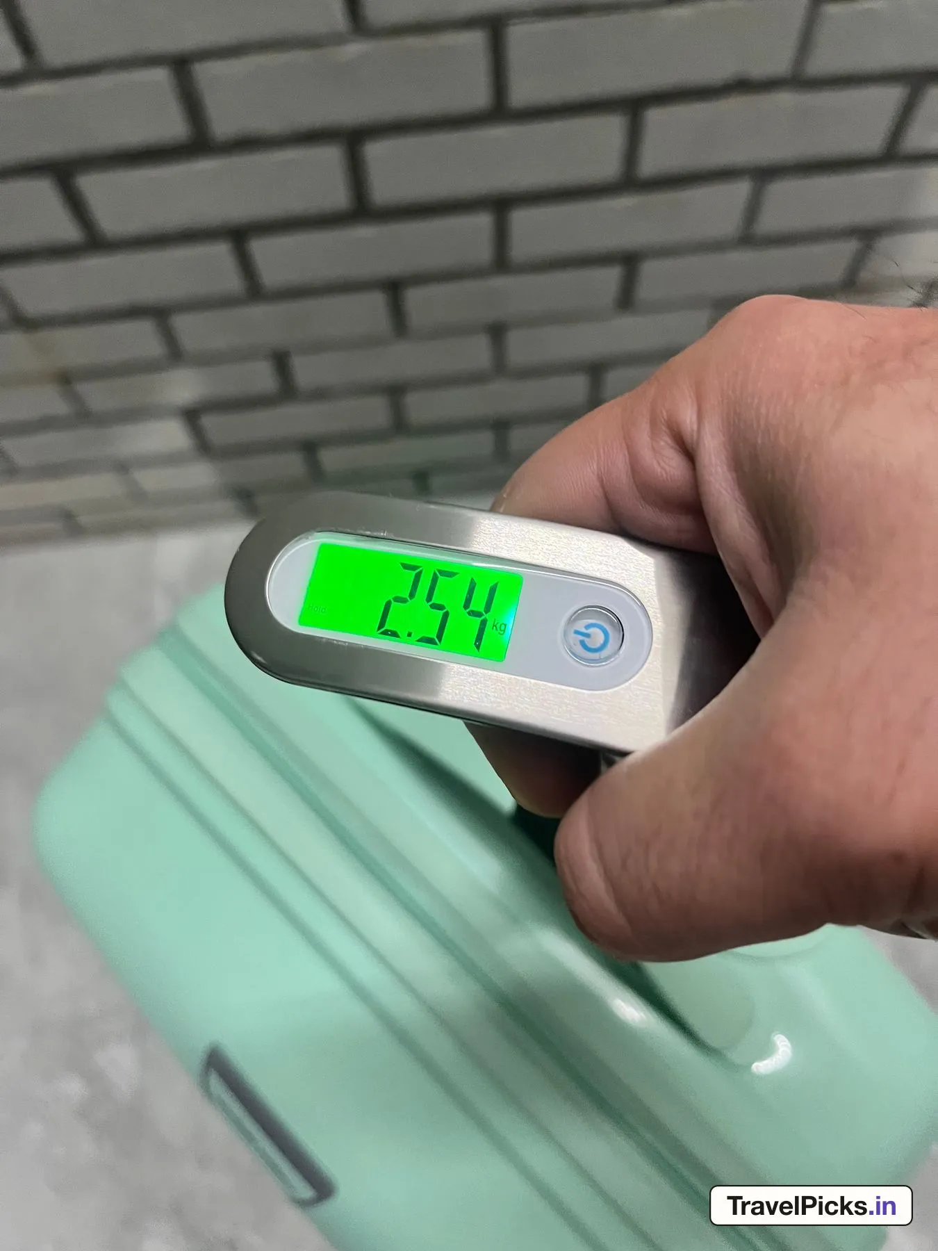 A hand-held digital luggage scale with a lit display reading 2.54 kg, the mint case hanging below it.